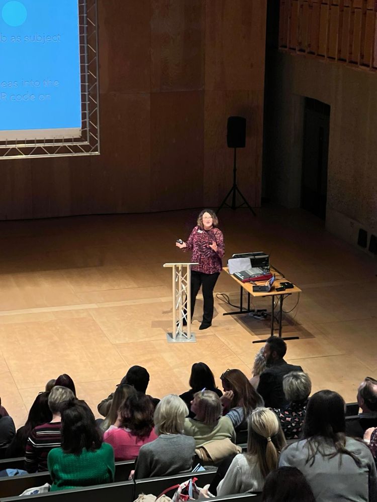 A female presenter on stage wearing black trousers and a red and purple top. 