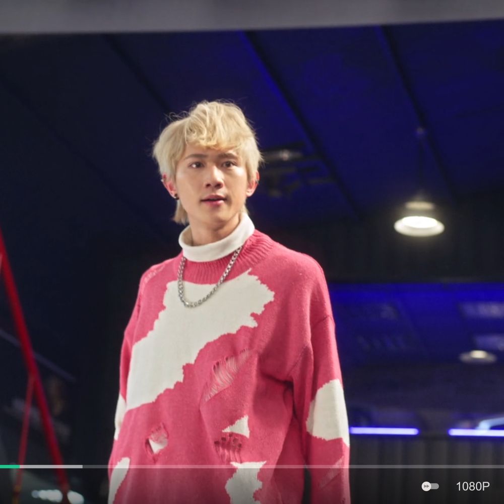 a screenshot of Ai Di, a character from the show Kiseki: Dear to Me. He has dyed blond hair, a silver chain necklace, and a bright pink and white sweater, which is fashionably splotched and fashionably ripped. He is standing in an upscale garage and has a look of frustration on his face.