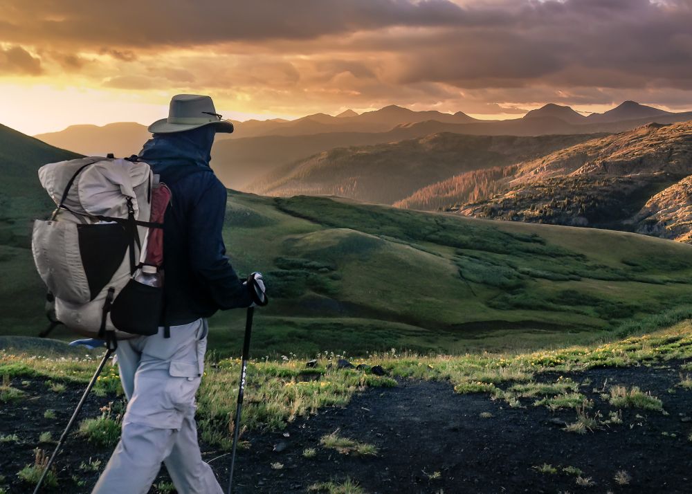 The image is of a Colorado Trail hiker on the trail in the early morning light, with mountains and a stormy sky in the background.