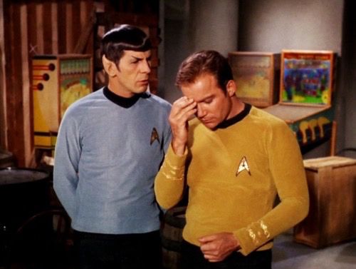 spock and jim kirk from star trek standing next to each other, spock is wearing a blue uniform and kirk is wearing a yellow one, spock is telling him something and kirk looks tired, he’s holding his head as if it’s hurting him