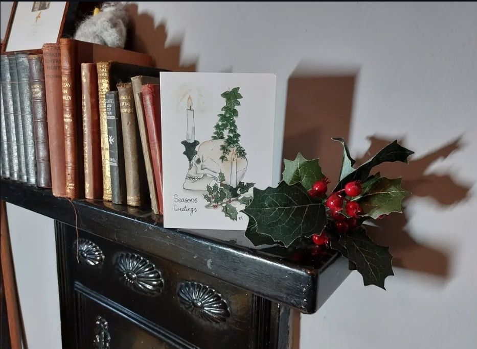 A Gothic Christmas scene: a black painted Edwardian fireplace against a white wall. One half of the mantle is filled with antique books in shades of navy db's brown leather. Next to the books is a greetings card and sprigs of faux holy and berries. The card depicts a watercolour illustration of a skull in profile with a tall candle, trailing ivy abs green holly and the phrase "seasons greetings".