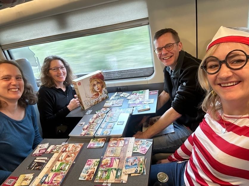 Donna and Matt Leacock, Elizabeth Hargrave, and Danielle Reynolds playing the boardgame Bohemians on a train