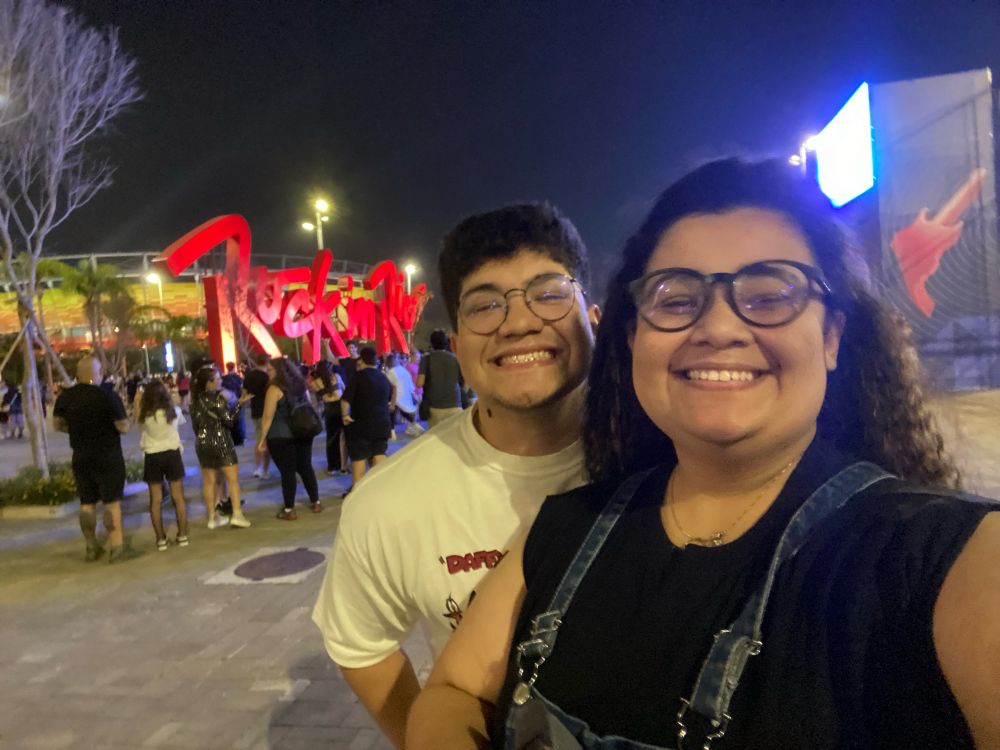 Foto de uma mulher de cabelo cacheado e plusa preta e um homem de blusa branca com a placa do Rock in Rio