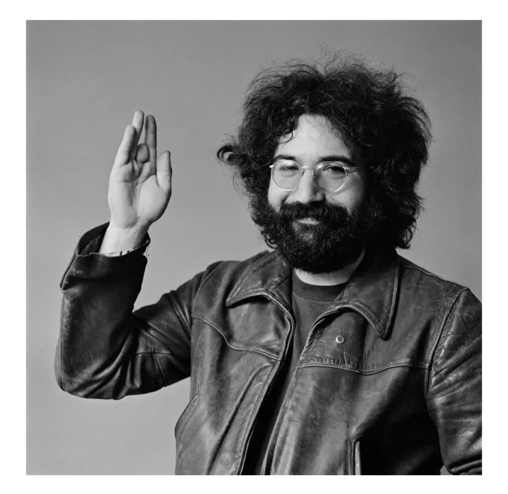 A man (Jerry Garcia) with bushy black hair and beard wearing a leather jacket with his right hand raised. 