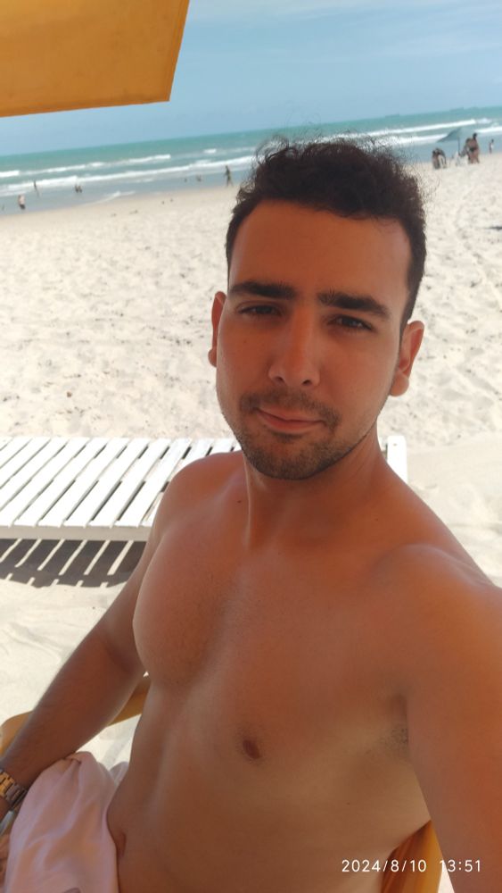 Foto de Lincon, na praia do Futuro - Fortaleza (CE). Em primeiro plano, sentado, na areia, com o mar ao fundo e o céu azul.