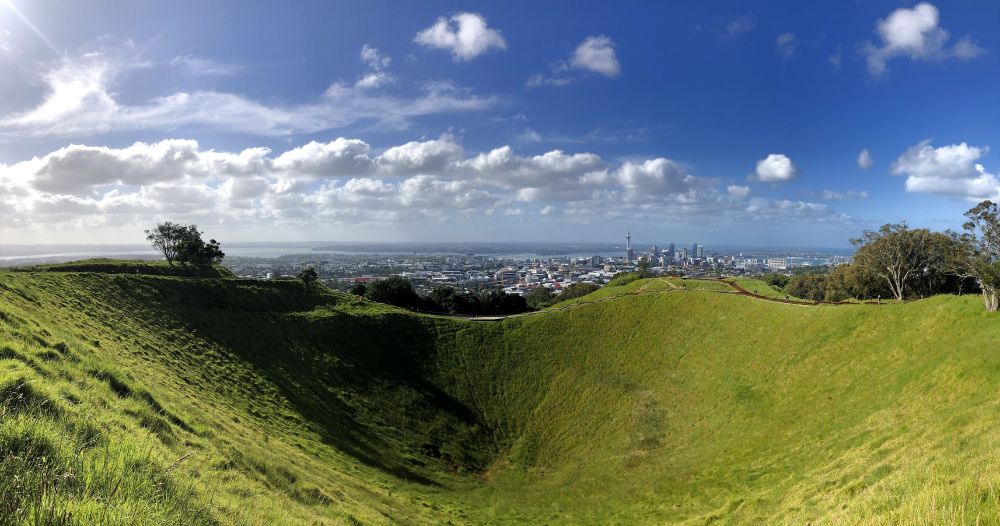 Picture of a large volcano crater, covered in grass. Bright blue sky with Auckland city skyline in the background and water in the distance. 