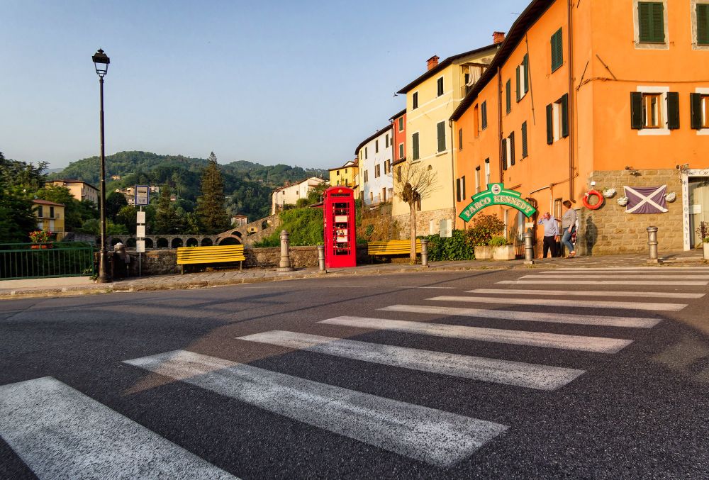 Colour photo of an Italian pedestrian crossing with orange and yellow painted buildings and wooded hills behind . There is a red telephone box on the other side of the road.

Olympus E-M10MarkII
LUMIX G VARIO 12-32/F3.5-5.6
@ƒ/6.3 12.0 mm 1/250s ISO200