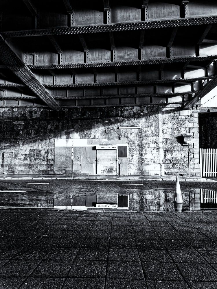 Black & white photo of a shuttered entrance with "C'mon in" above the door. The entrance is in a stone wall under a iron bridge, the roadway has a large puddle reflecting the entrance.
Olympus E-5, Olympus 12-60mm lens
@ƒ/2.8 12.0 mm 1/80s ISO160