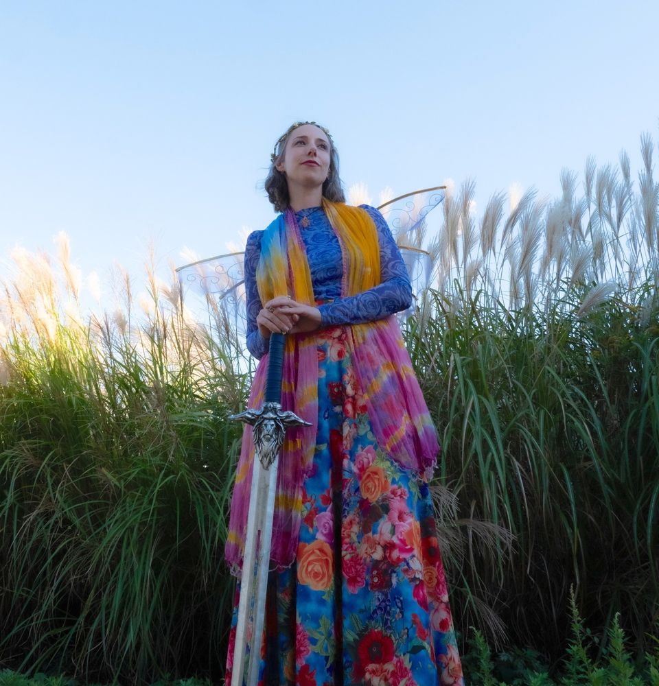 Stephanie, a white femme, stands against a backdrop of tall green plants and pale blue sky. She is dressed in colorful floral attire with fairy wings. She looks purposefully into the distance while her hands rest on a silver sword with a lion motif.  
