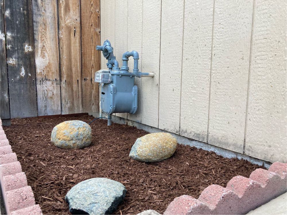 Area of mulch against side of house with brick trim and decorative rocks. A gas meter is on the side of the house and a fence is at the back of the photo. 