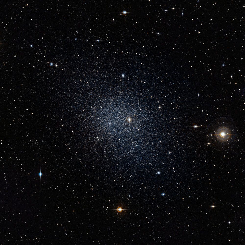 A deep-space image of the Fornax Dwarf Galaxy, showing a dense concentration of faint stars at the center surrounded by a sparse field of individual stars. The galaxy appears as a hazy, diffuse glow against the black background of space, with a few bright foreground stars creating starburst effects.