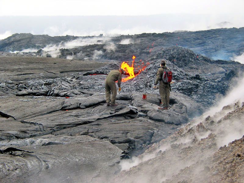 Two volcanologists in protective gear collect lava samples from an active volcanic vent ok Kilauea Volcano in 2004, with glowing molten lava emerging from a vent and steam rising from the surrounding hardened black lava field.
