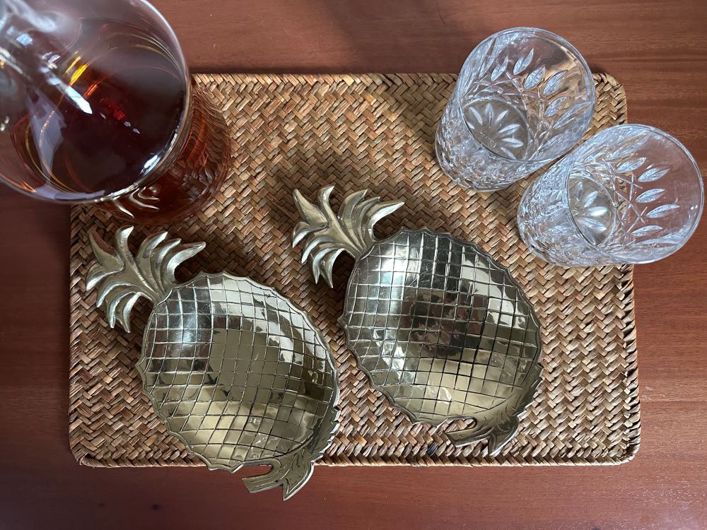 Picture of two brass pineapple dishes on a rattan placemat. There’s also two cut crystal glasses and a whiskey decanter.