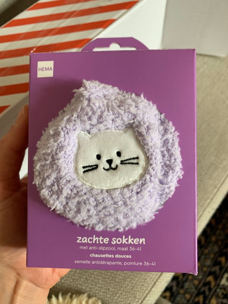 hema „zachte sokken“ (weiche flauschige socken) fliederfarbener flausch mit samtigem katzenkopfmotiv schaut aus einer lilafarbenen viereckigen verpackung hervor, diese befindet wird von meiner hand gehalten 