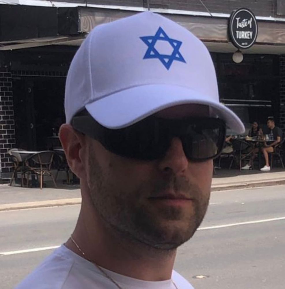 a man wearing dark sunglasses and a white cap with a large blue star of david symbol on it 