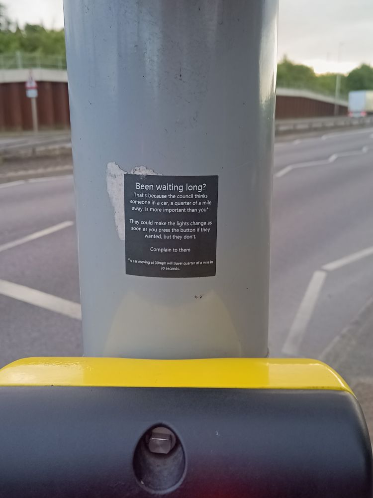 A sticker above a pedestrian beg button by a busy road. It says: "Been waiting long? That's because the council thinks someone in a car a quarter of a mile away is more important than you.

They could make the lights change as soon as you press the button if they wanted to, but they don't.

Complain to them." 