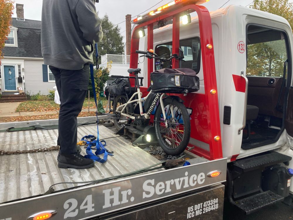 Cargo e-bike on the front of the bed of a tow truck that can probably handle two large cars. The driver is tying it on with cinch straps 