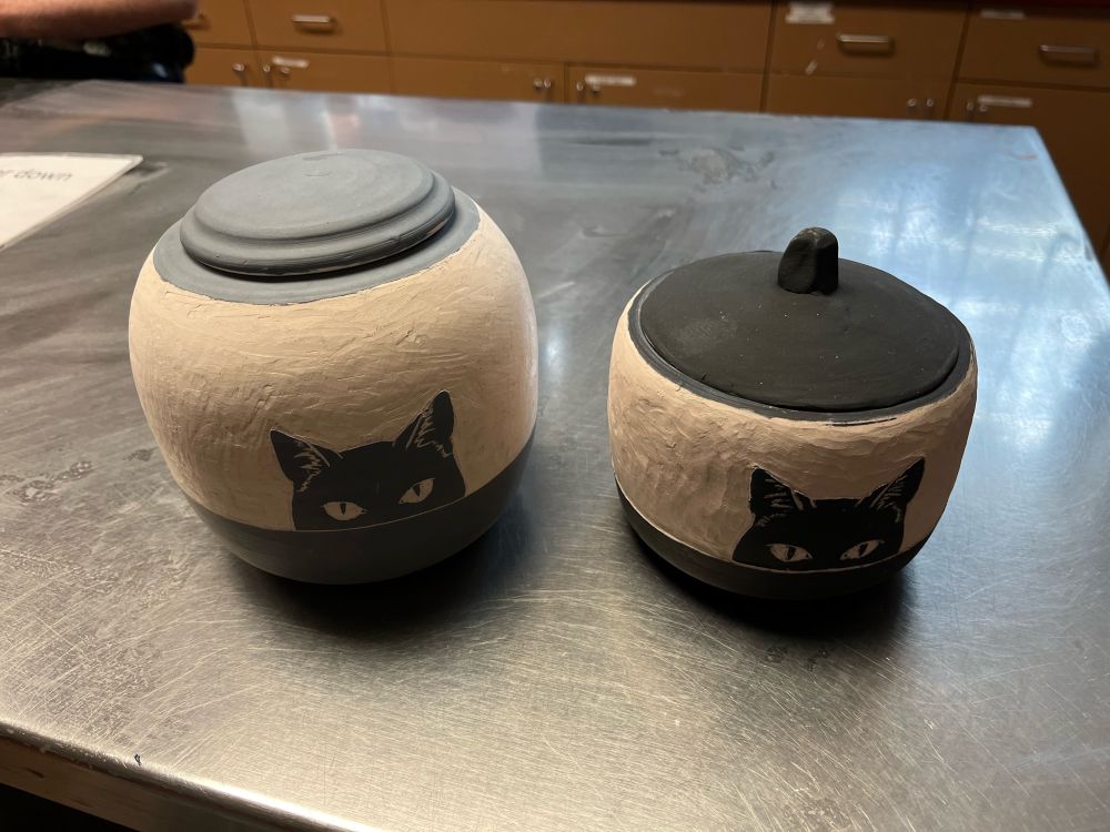 Two pots that have been bisque fired, one with a grey top and lid, one with a black top and lid. The image of cat eyes peering over the edge of a table/counter are carved into each pot.