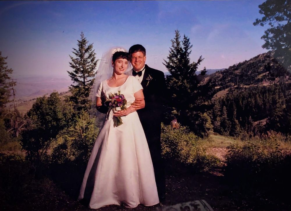 A bride and groom standing together, smiling. Cream colored long satin dress, with short sleeves and fingertip veil behind her head. Groom is in a black tuxedo. Bride is holding a bouquet of blue and burgundy flowers. They are standing on a hill in the Boise mountains with a brilliant blue sky and tall pine trees.