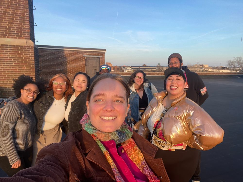 7 people, with golden hour sunset sun on their faces smile at the camera together. In the background, the roof of a high rise building and the city scape of Detroit. 