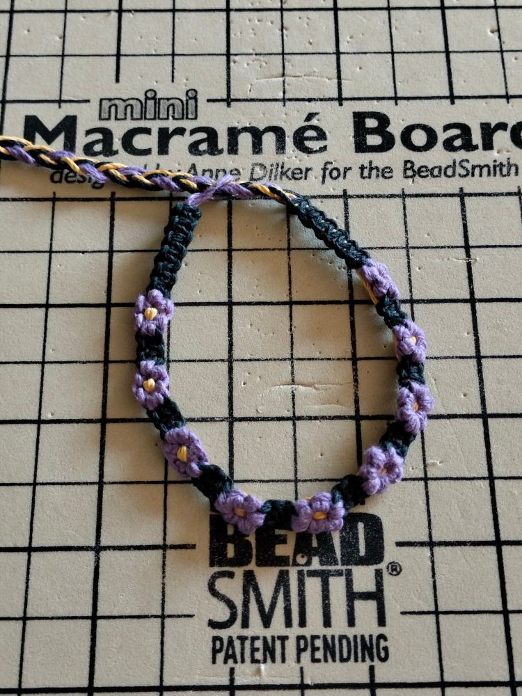 A woven bracelet with flowers with purple petals and a yellow centre, with black square knots in-between, resting on a beige foam board with black lines and the text "mini macrame board".