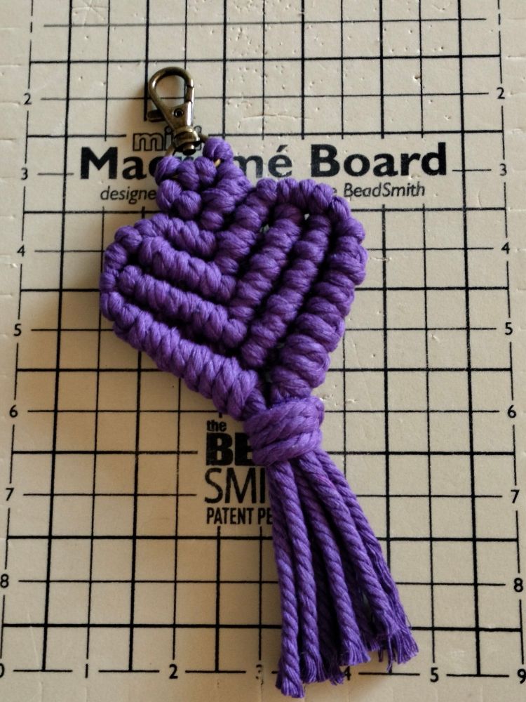 A purple macrame heart with a silver clip and purple tassels, resting on a beige foam board with black lines and the text "mini macrame board".