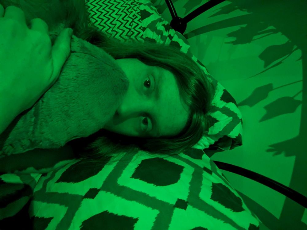 Willow in bed cuddling her blahaj. The lighting is green and she is holding the shark up to her face