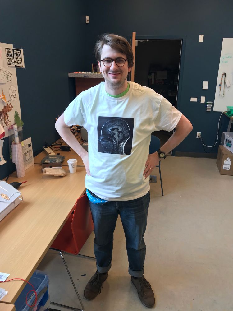 A white man in his early 30s stands smiling with his hands on his hips. He’s wearing a white t-shirt, on which is printed a black and white image of an MRI brain scan.