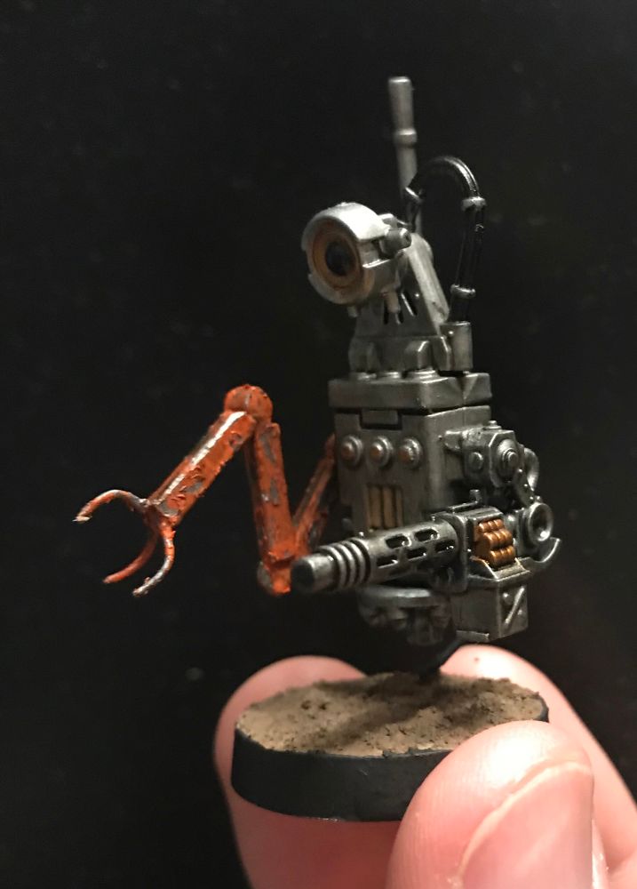 A miniature robot that appears to have been built from a variety of mismatched parts. It hovers (it’s attached to the base with a black wire), and it has a long antenna on its back. Its right arm is some sort of industrial grabber, and the orange paint is peeling off. Its left arm is a large machine gun. The robot has one large eye on top.