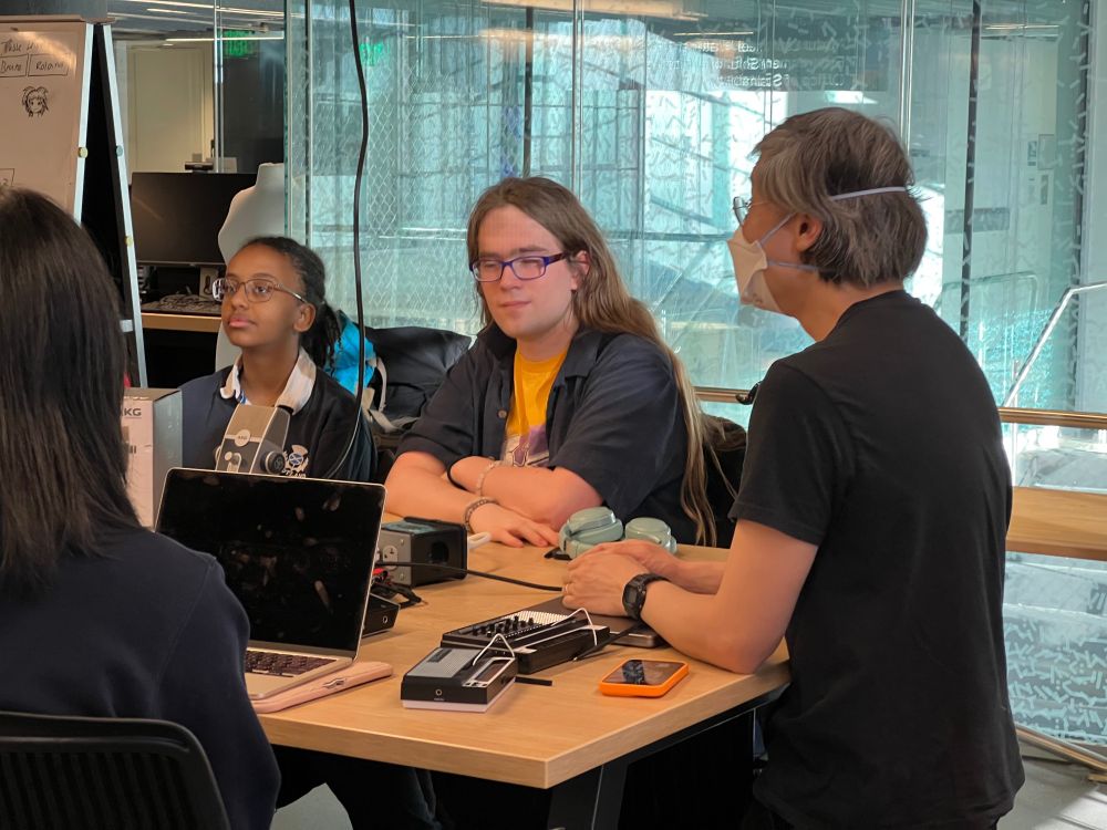 Philip tan (right) at a table talking to students (3 pictured) about playing with musical toys. A couple stylophones are on the table.