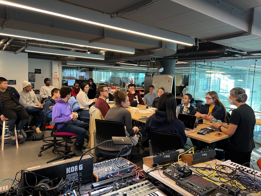 View of the full Voxel Lab with a bunch of synthesizers in the foreground (mostly Korg equipment) and a table with 15+ students around it listening to Philip talk about playing with musical toys.
