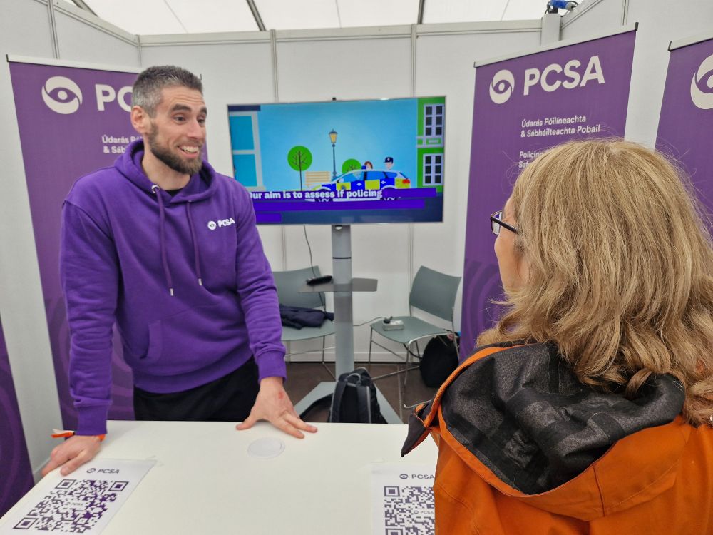 PCSA staff member chatting to member of the public at counter with PCSA purple branding in the background including television with PCSA branding playing 