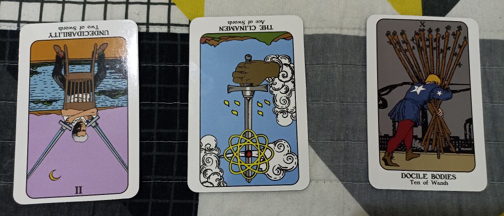 Tiragem de três cartas do tarô, com baralho "Philisophers' Tarot". À esquerda, "Undecidability" (2 de espadas) de cabeça para baixo. No centro, "The Clinamen" (às de espadas), também de cabeça para baixo. Na direita, "Docile bodies" (dez de paus).