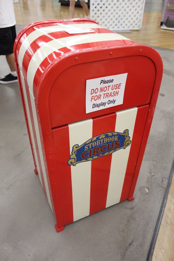 A decorative trash can designed to resemble circus stripes, predominantly red and white. It features a sign that says "Please DO NOT USE FOR TRASH" and notes that it is for display only. The trash can is set in an indoor location with a wooden floor.