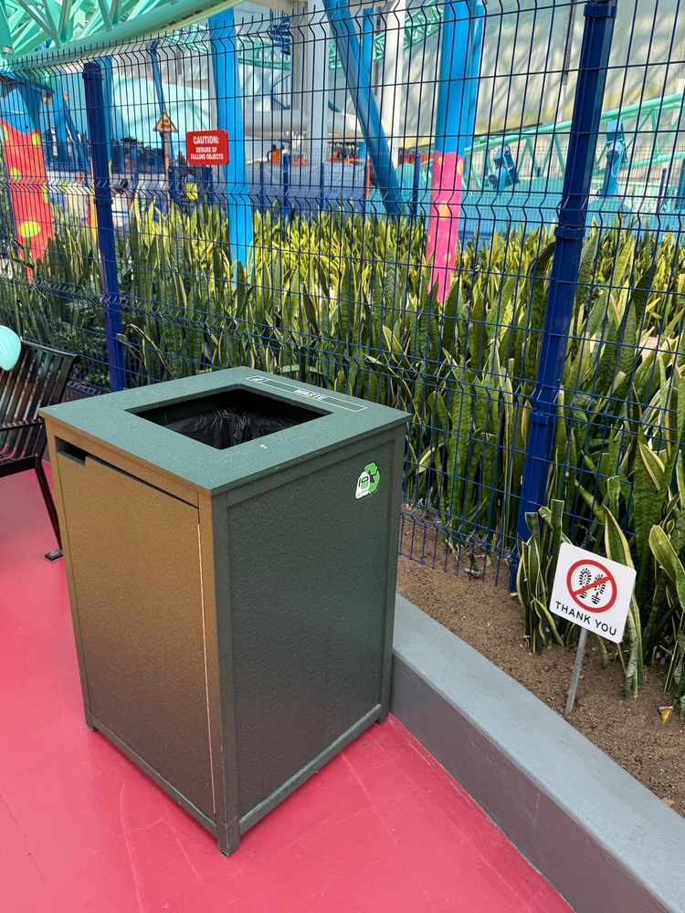 A green trash bin with a closed lid is positioned on a pink surface. Behind it is a fence with a caution sign and an area of plants. A small sign nearby asks for no littering.