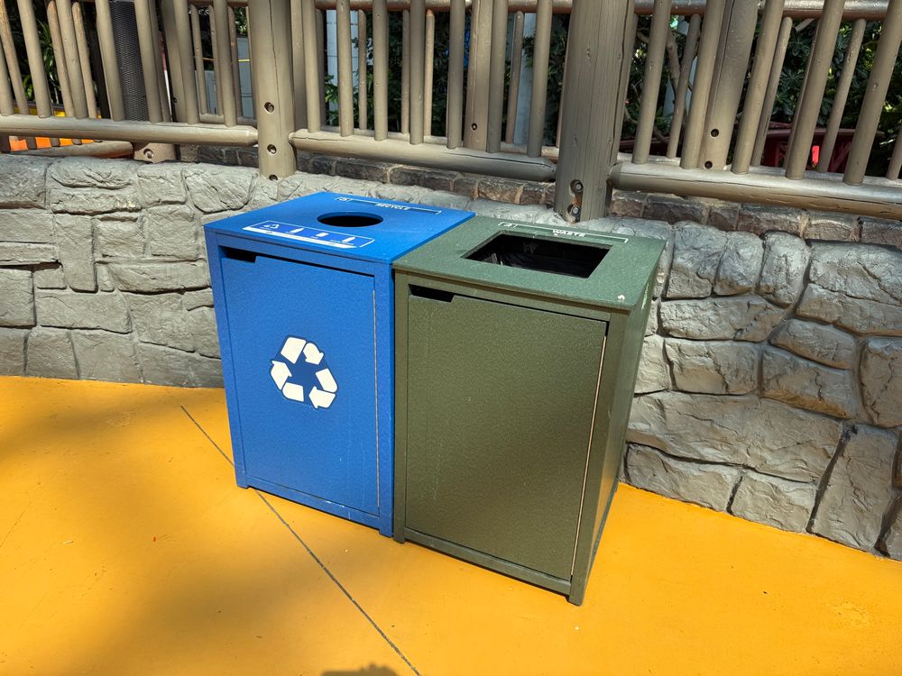 Very wise to read the ALT Text. Here's an extra hint: Note the shadows on the ground.  

Back to the ALT text: Two trash cans placed side by side; one is blue with a recycling symbol, and the other is green, both set against a stone wall and yellow floor.