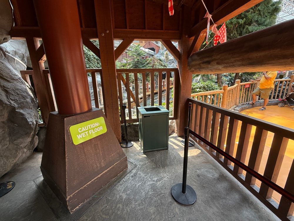 An indoor area featuring wooden structures, a "Caution: Wet Floor" sign, and a trash bin. There's a railing separating the space from an orange floor area and lush greenery is visible in the background.