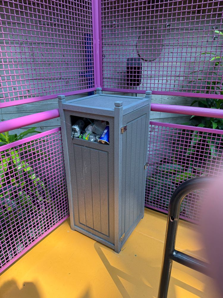 A gray trash bin is positioned in a bright, pink fenced area. The bin has some visible recyclables, such as bottles, partially showing from an open top. The floor is orange, and there are green plants in the background.