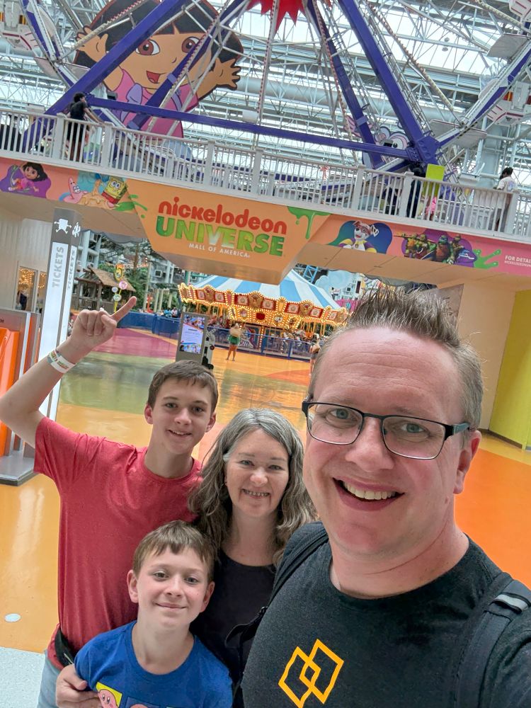 A family of four poses for a selfie in Nickelodeon Universe at the Mall of America. In the background, colorful rides and a large sign for Nickelodeon Universe can be seen. The area has a bright, fun atmosphere with an indoor amusement park. 