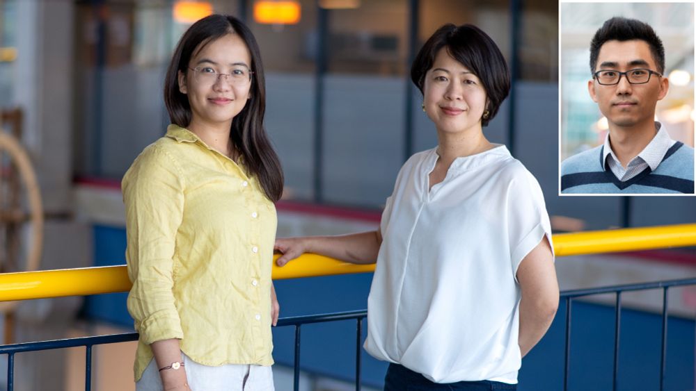 Two women stand together near a yellow railing in a modern indoor space. A smaller image of a man is inset in the corner.