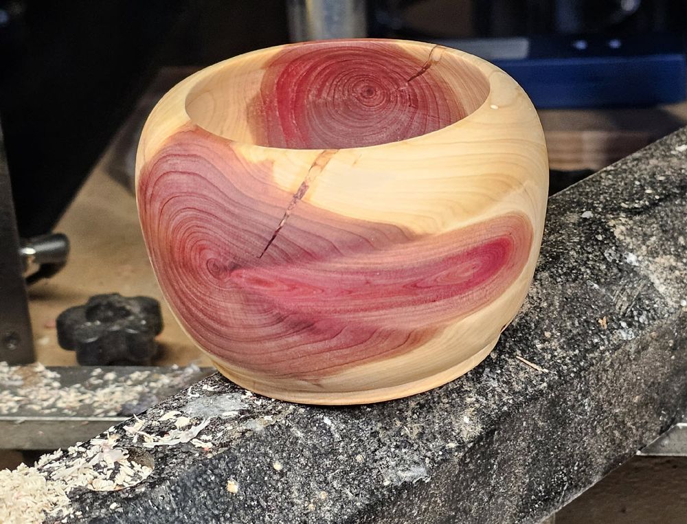 Photo of a small 3x3 inch cedar bowl sitting on the bed of a wood lathe. The bowl is cream colored with burgundy colored heartwood. The limb orientation was perpendicular to the bowl so the heartwood is on opposite sides of the bowl. The limb was cracked and the 1/8 inch crack is visible from the top of the bowl to halfway down and slanted to the left. It was filled with twigs and sawdust and bound with super glue. On finishing, the twigs show as cross sections running down the crack. 