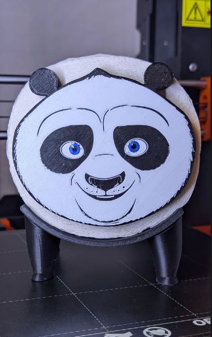 Toilet paper roll on its side sits in a holder that had 4 stubby black legs and paws. A panda face is 3D printed and held on the side of the roll by the toilet paper core.
