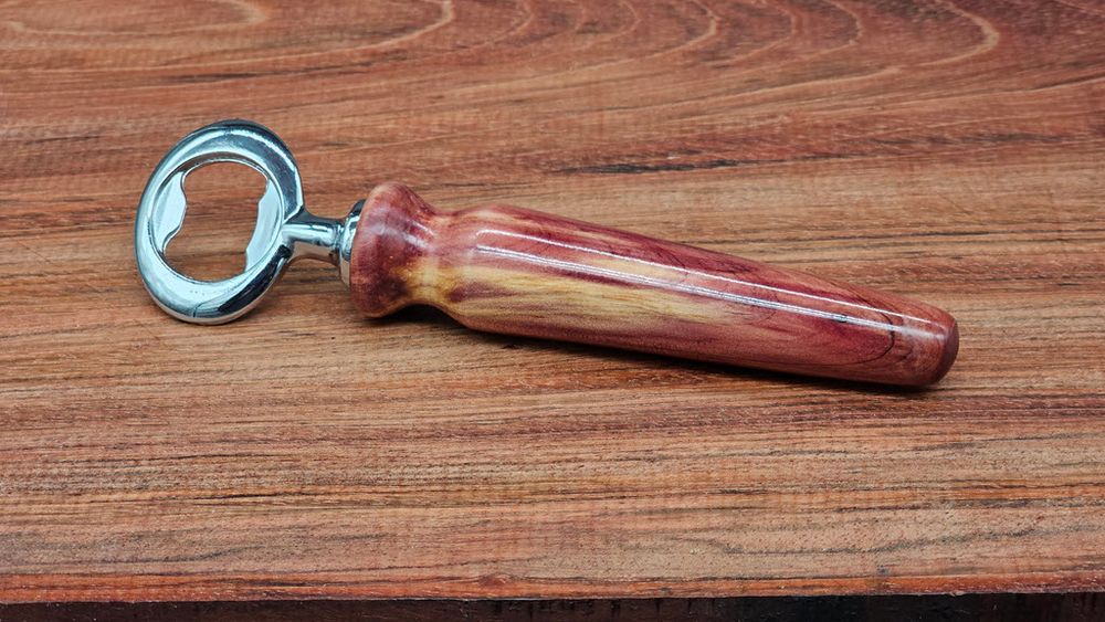 Photo of a bottle opener with a cedar handle resting on a natural wood surface. The handle shows a mix of yellow-cream colored sapwood and the bright read heartwood for which cedar is known. The handle has a shiny gloss finish.