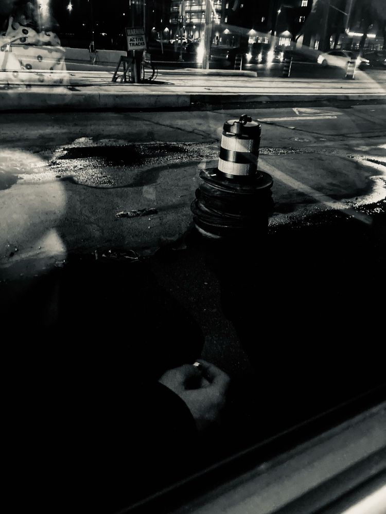 Heavily edited photo from a window bus seat, partially wet ground, a pylon in the middle of the road near tram tracks. Photo is filtered with a silver tone setting and with other settings adjustments.