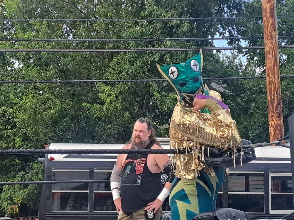 Shot of a wrestling ring with a frog costume wrestler posing on the corner and a metal head wrestler looming behind him 
