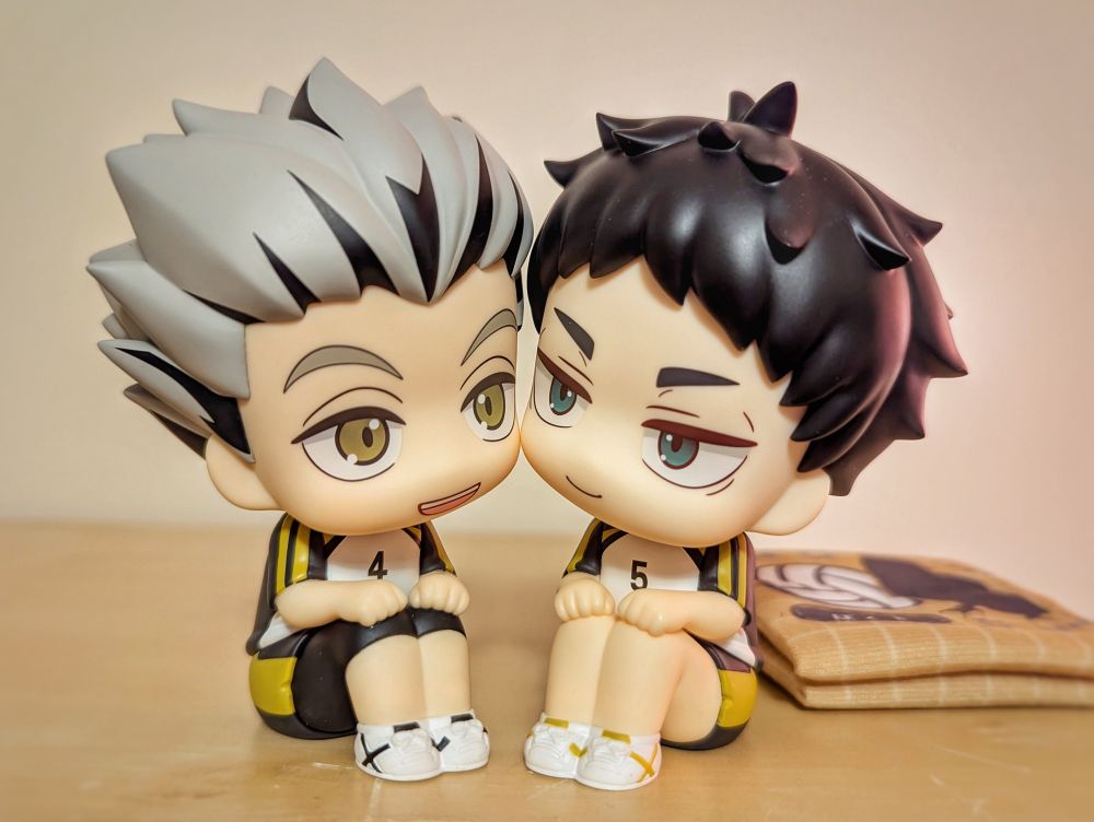 Photograph of chibi figurines from the "look up" series of Bokuto and Akaashi from Haikyuu in their Fukurodani uniforms. Here they are sitting next to each other positioned to look like their cheeks are touching. 