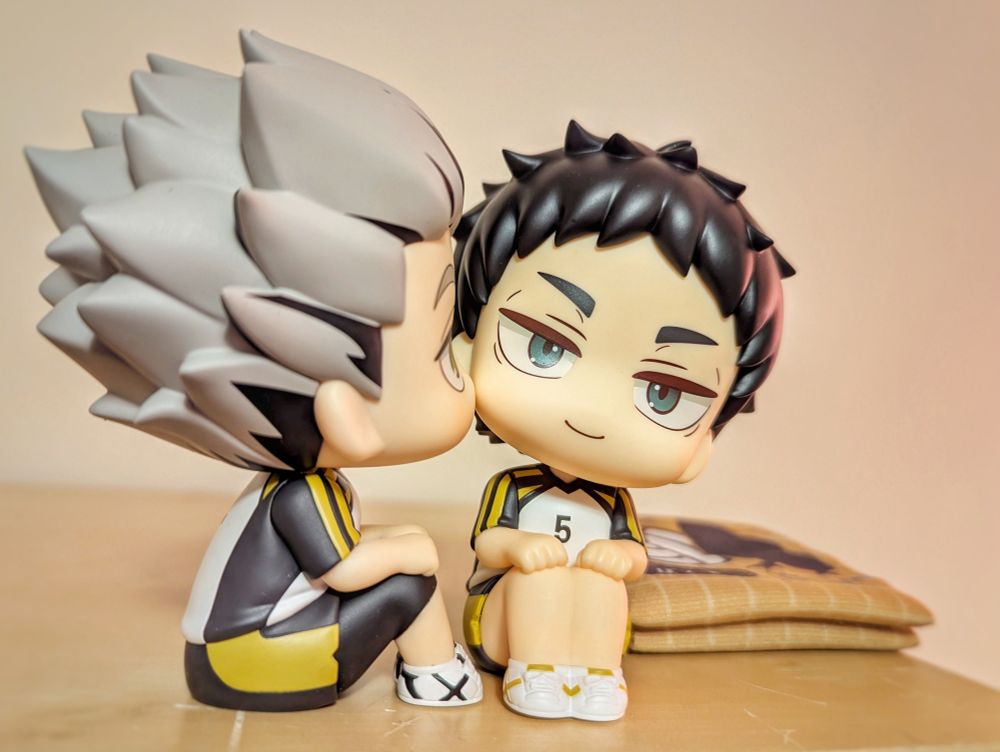 Photograph of chibi figurines from the "look up" series of Bokuto and Akaashi from Haikyuu in their Fukurodani uniforms. Here they are sitting next to each other positioned to look like Bokuto  is kissing Akaashi's cheek.