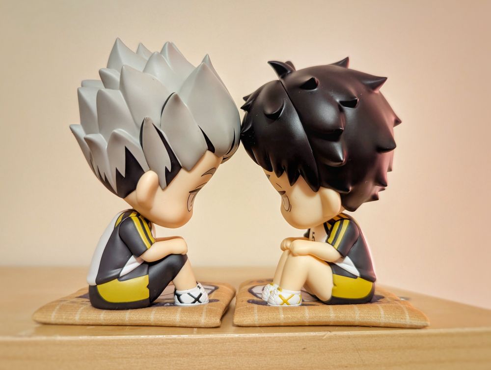 Photograph of chibi figurines from the "look up" series of Bokuto and Akaashi from Haikyuu in their Fukurodani uniforms. Here they are sitting on tiny floor cushions and their heads are angled so they look like they're pressing their foreheads together. 