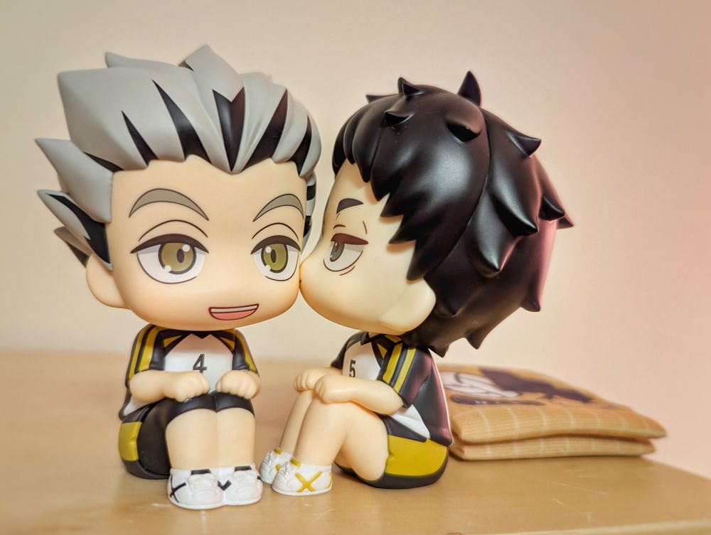 Photograph of chibi figurines from the "look up" series of Bokuto and Akaashi from Haikyuu in their Fukurodani uniforms. Here they are sitting next to each other positioned to look like Akaashi is kissing Bokuto's cheek.