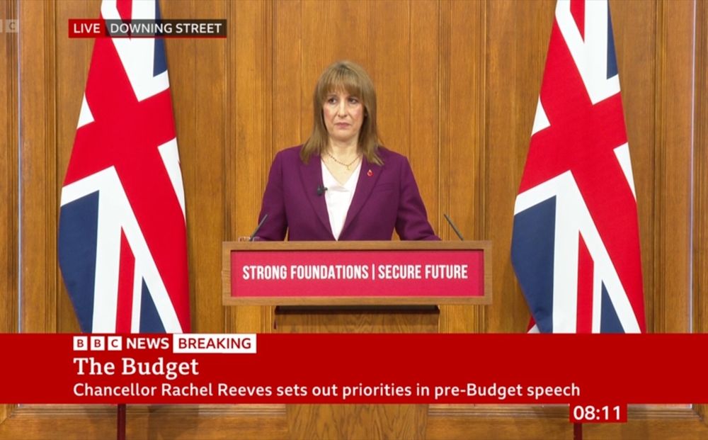 Rachel Reeves at a lectern in front of a wood-panelled wall and two huge British flags. The lectern has a red sign: "Strong Foundations / Secure Future". A red bbc news chyron reads "The Budget"